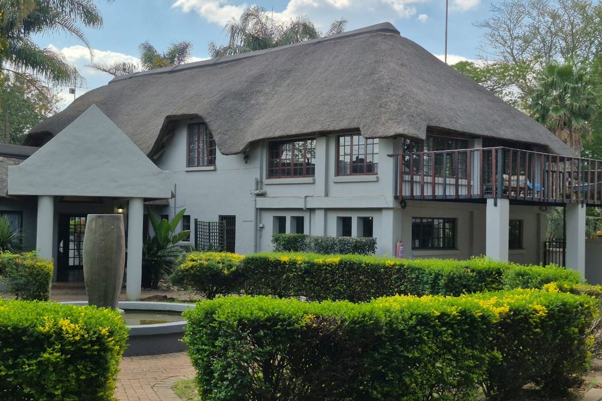 A rear view of a traditional thatched-roof house featuring a balcony, landscaped garden, paved pathways, and a decorative water feature.