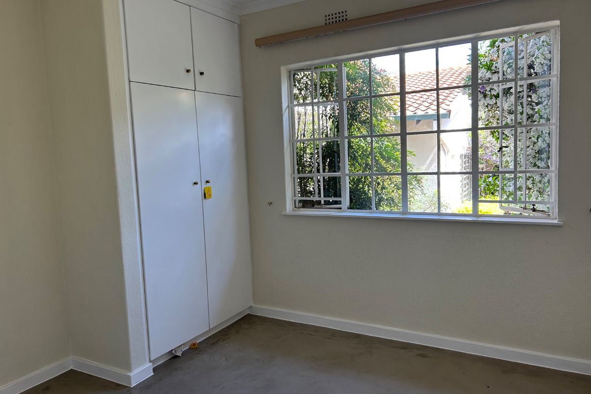 An empty bedroom featuring built-in cupboards and a large steel-framed window with security bars, offering a view of the garden.