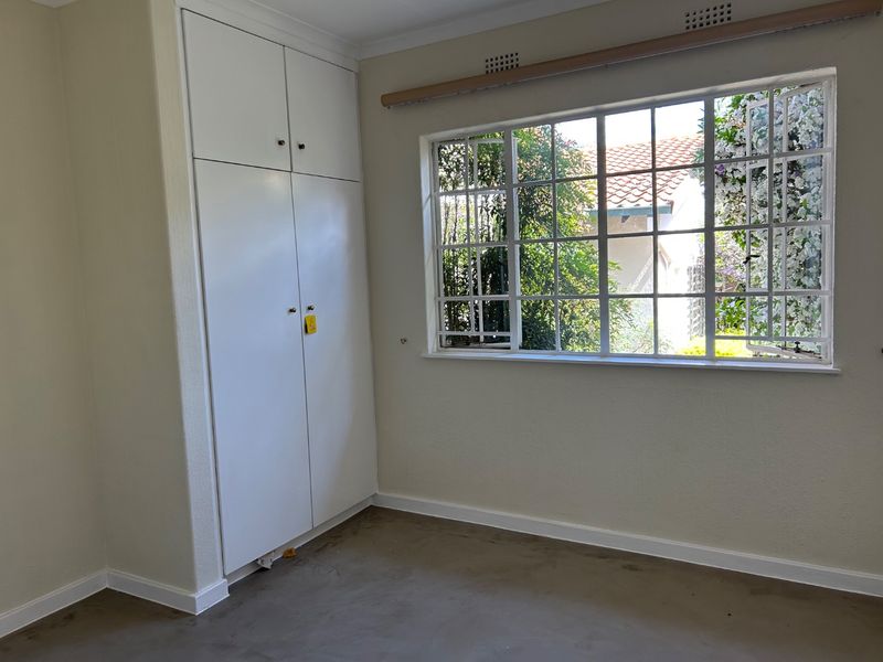 An empty bedroom featuring built-in cupboards and a large steel-framed window with security bars, offering a view of the garden.