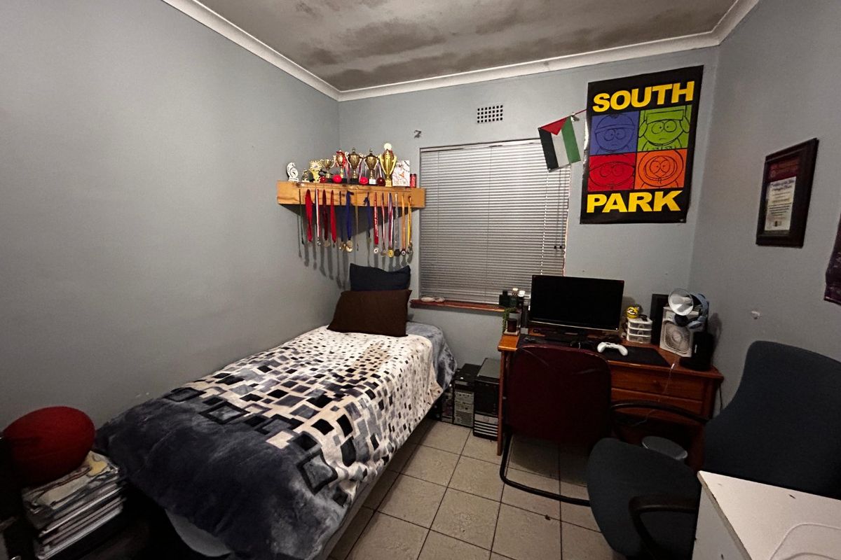 A compact bedroom featuring tiled flooring, venetian blinds, and a wall-mounted shelf. The ceiling shows visible signs of water staining.