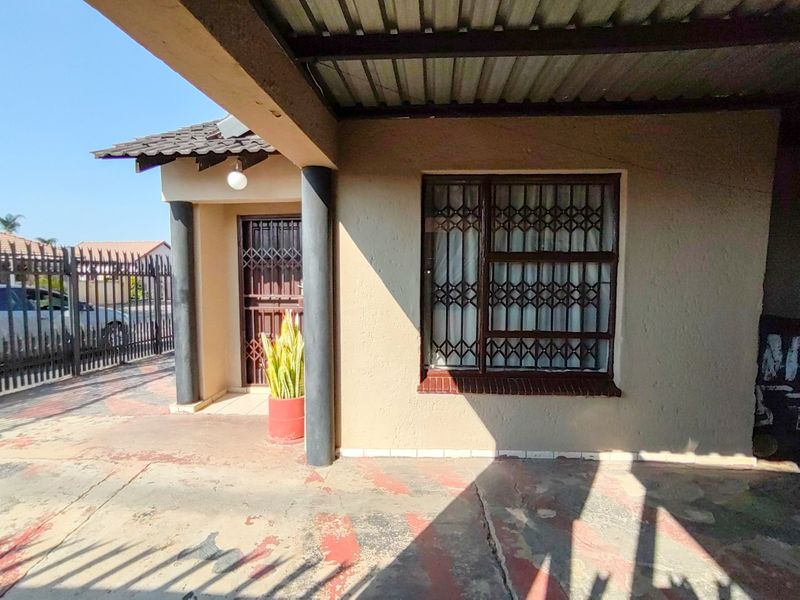 Front entrance area featuring a covered carport, security gates, and security bars on windows, with a concrete driveway showing signs of wear.