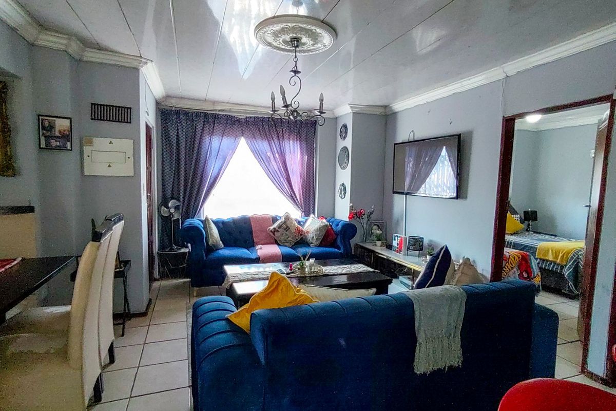 A living room featuring tiled flooring, decorative ceiling rose, and coved cornicing, with an adjacent bedroom visible through the doorway.