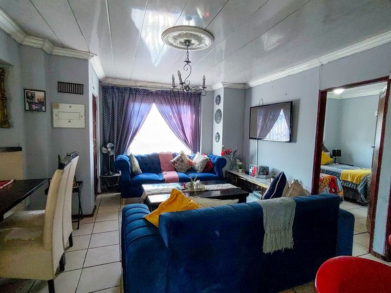 A living room featuring tiled flooring, decorative ceiling rose, and coved cornicing, with an adjacent bedroom visible through the doorway.