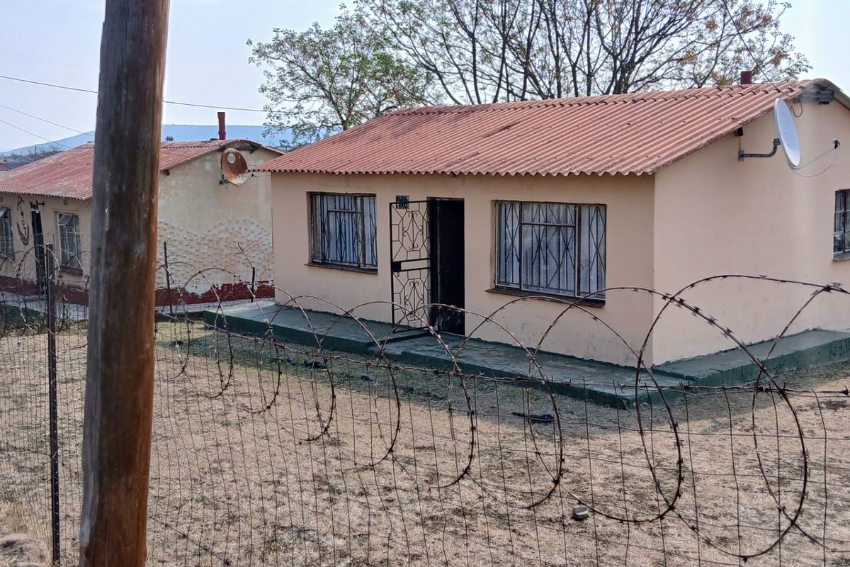 A detached single-story house featuring a corrugated metal roof, security gates, and burglar bars, enclosed by a wire fence with razor wire.