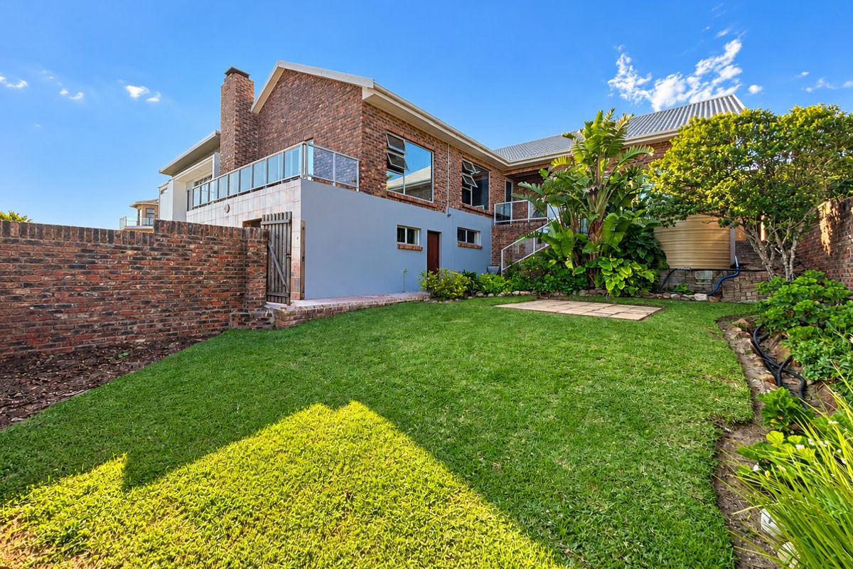 Rear view of a multi-level brick house featuring a landscaped garden, paved patio area, and a glass-railed balcony.