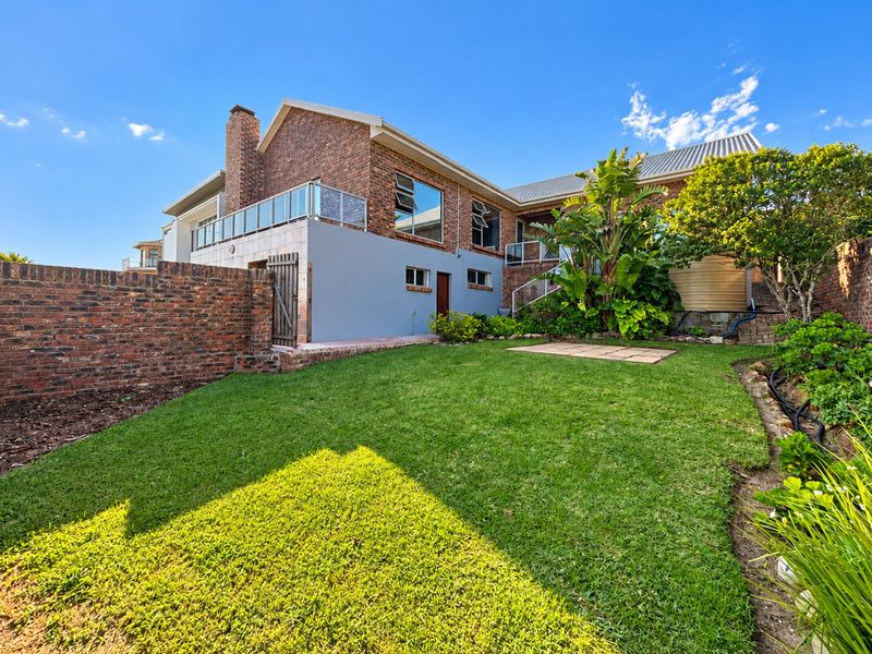 Rear view of a multi-level brick house featuring a landscaped garden, paved patio area, and a glass-railed balcony.