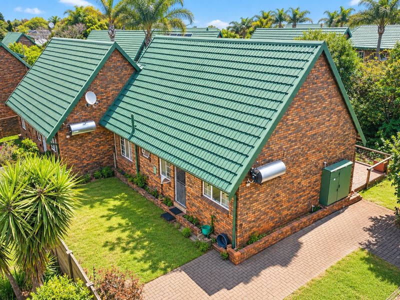 Aerial view of a brick townhouse featuring a green pitched tiled roof, solar geyser, private lawn, and paved driveway area.