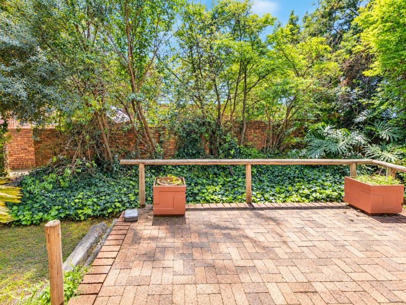 A private outdoor patio area featuring brick paving, a timber safety railing, and a mature garden backdrop with a brick boundary wall.