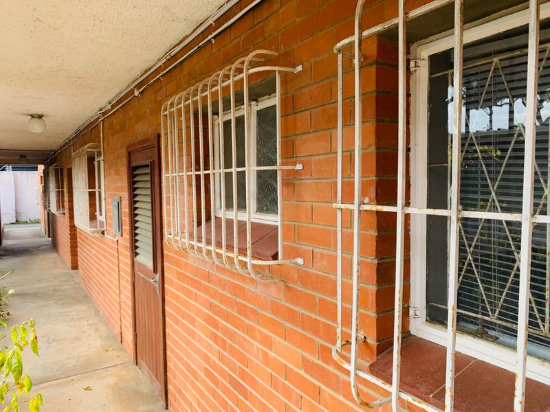 Exterior view of an apartment walkway featuring exposed brick walls, louvered doors, and security bars on windows showing signs of wear.