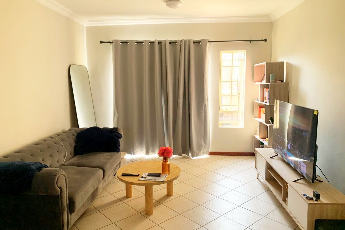 A well-lit living room featuring neutral tiled flooring, coving, and a window fitted with security bars and curtain rods.