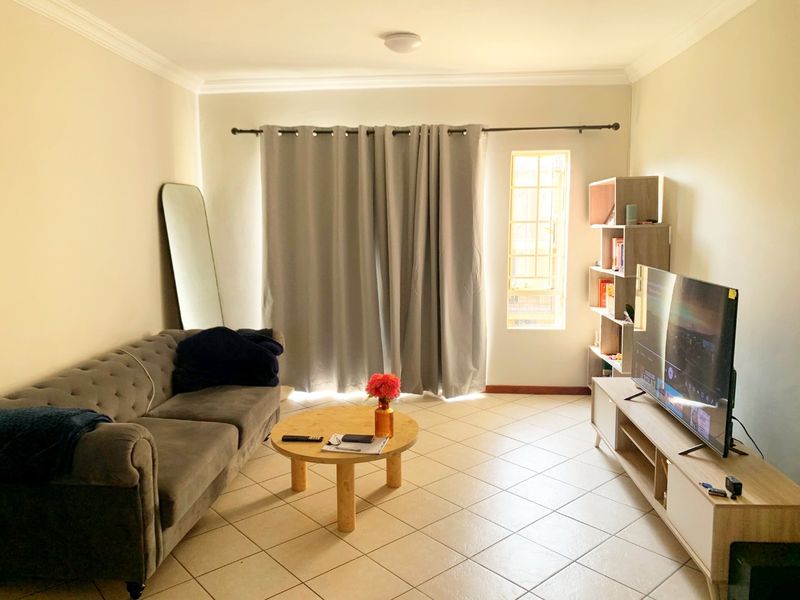 A well-lit living room featuring neutral tiled flooring, coving, and a window fitted with security bars and curtain rods.