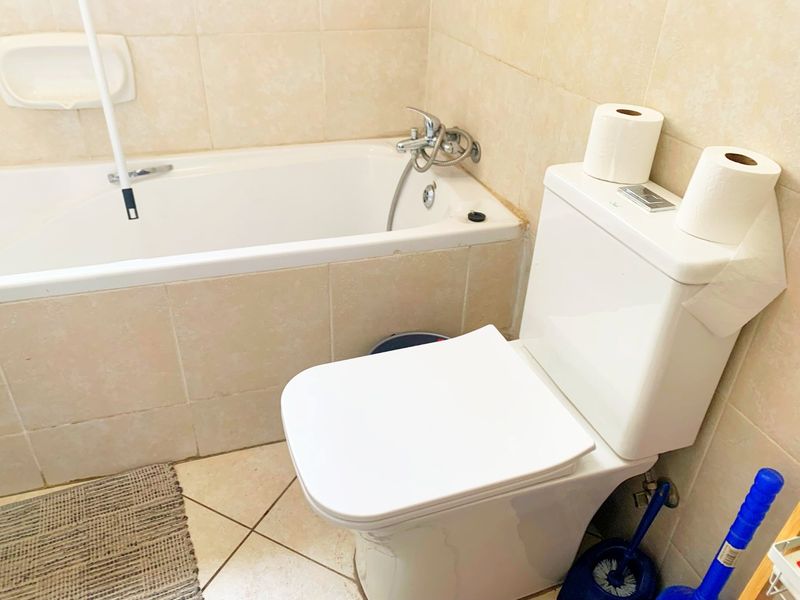 A functional bathroom featuring a built-in bathtub with shower mixer and a dual-flush toilet, finished with neutral ceramic wall and floor tiling.