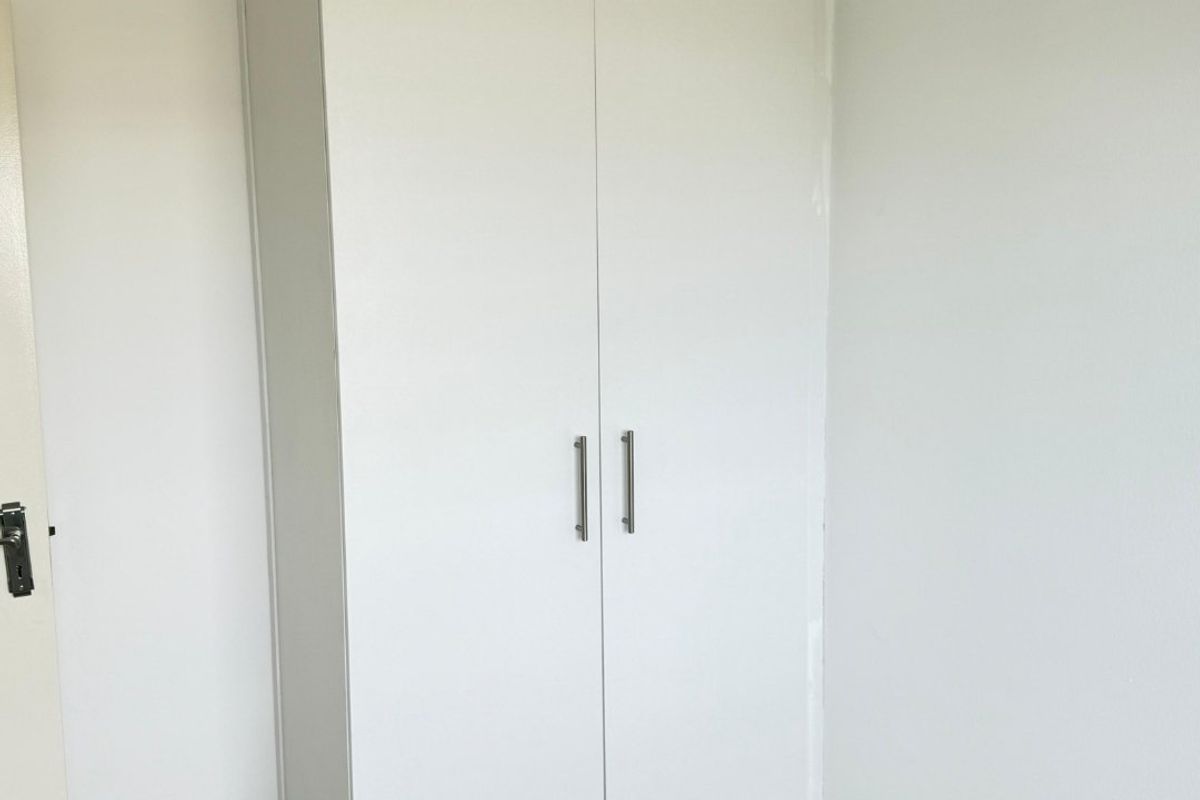 A clean, minimalist bedroom featuring a white built-in cupboard, neutral tiled flooring, and recessed ceiling lighting.