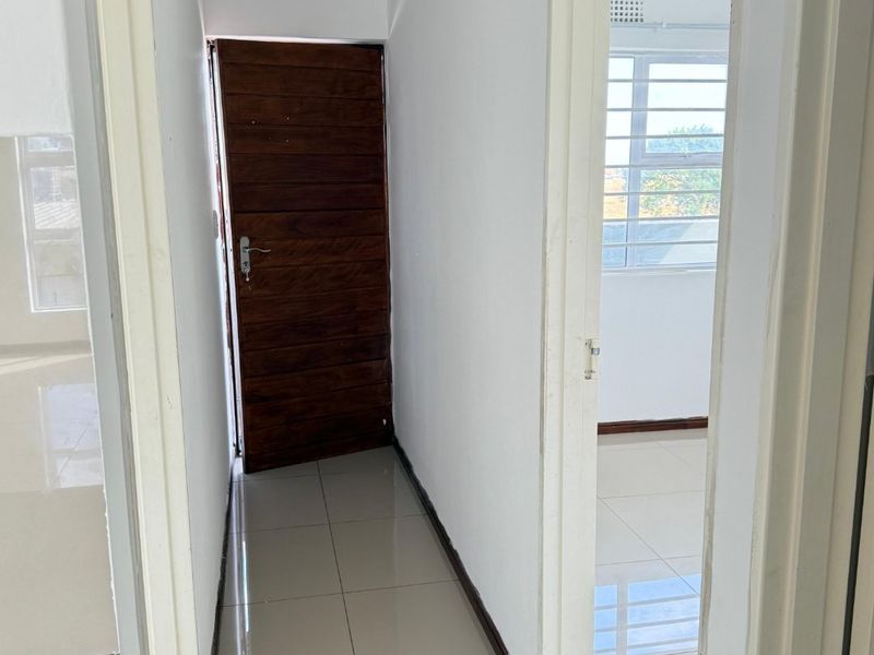 A clean, neutral hallway featuring tiled flooring, a solid timber door, and recessed ceiling lighting, providing access to adjacent rooms.
