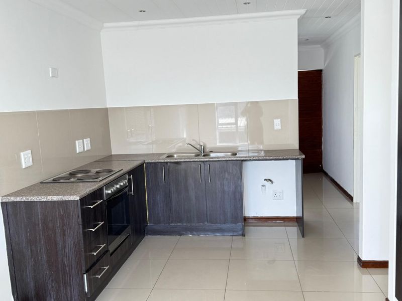 A clean, modern kitchen featuring dark wood cabinetry, granite countertops, integrated oven and hob, and neutral tiled flooring in an open-plan layout.