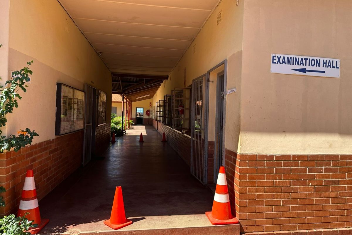 A covered outdoor hallway with brick base walls and security gates, leading towards an examination hall.