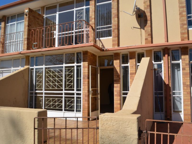 Front elevation of a townhouse featuring a ground floor patio, upper balcony, brick facade, and secure metal window grilles.