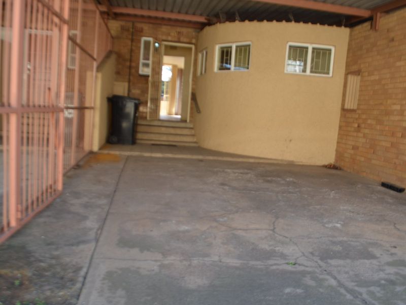A covered parking area or carport featuring a concrete floor, brick walls, and metal security gate access to the main entrance.