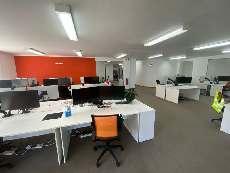 Open-plan commercial office space featuring neutral carpeting, bright fluorescent lighting, and a vibrant orange accent wall.