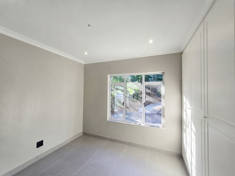 A bright, empty bedroom featuring neutral walls, tiled flooring, built-in cupboards, and recessed downlights, offering a clean and modern space.