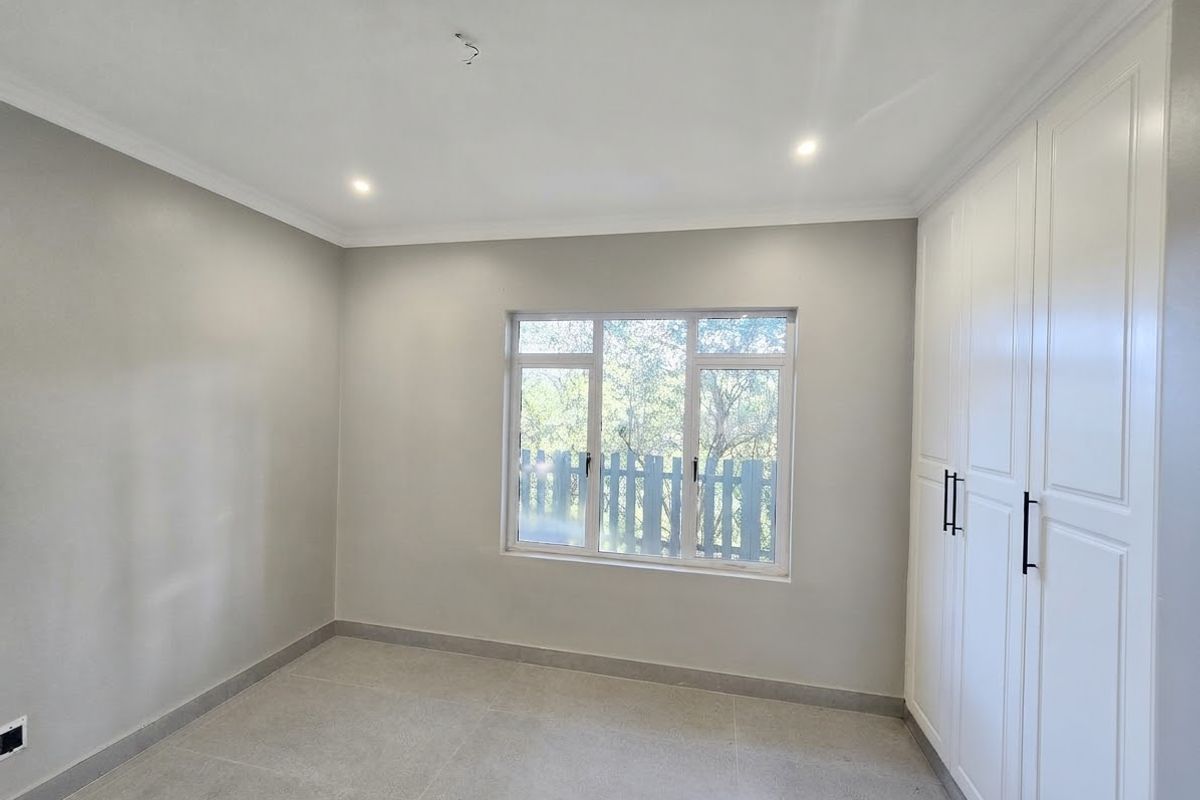 A bright, modern bedroom featuring neutral tiled flooring, built-in white cabinetry, and large aluminium windows allowing for ample natural light.