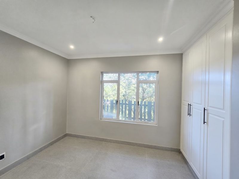 A bright, modern bedroom featuring neutral tiled flooring, built-in white cabinetry, and large aluminium windows allowing for ample natural light.