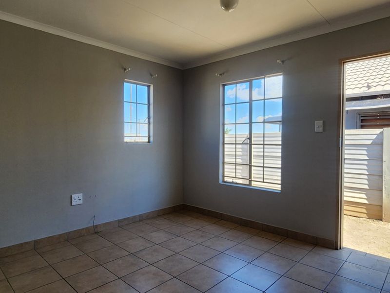A clean, empty bedroom featuring neutral wall tones, tiled flooring, and natural light through grid-pattern windows with direct access to the exterior.