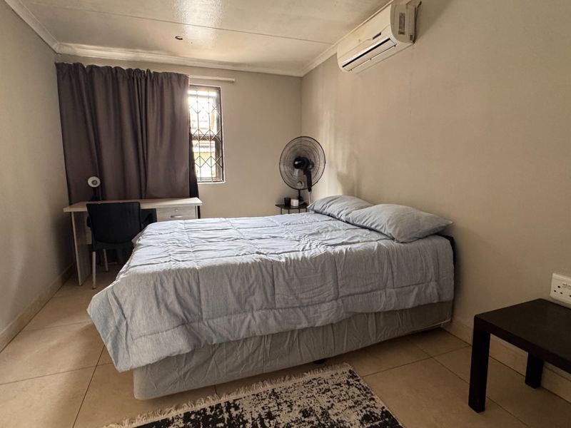 A functional bedroom featuring tiled flooring, an integrated air conditioning unit, and a window with security bars.