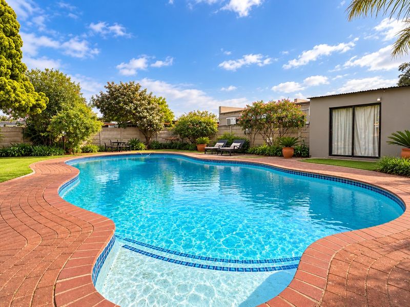 A well-maintained swimming pool area featuring brick coping, lush landscaping, and a clear view of the rear garden and adjacent structure.