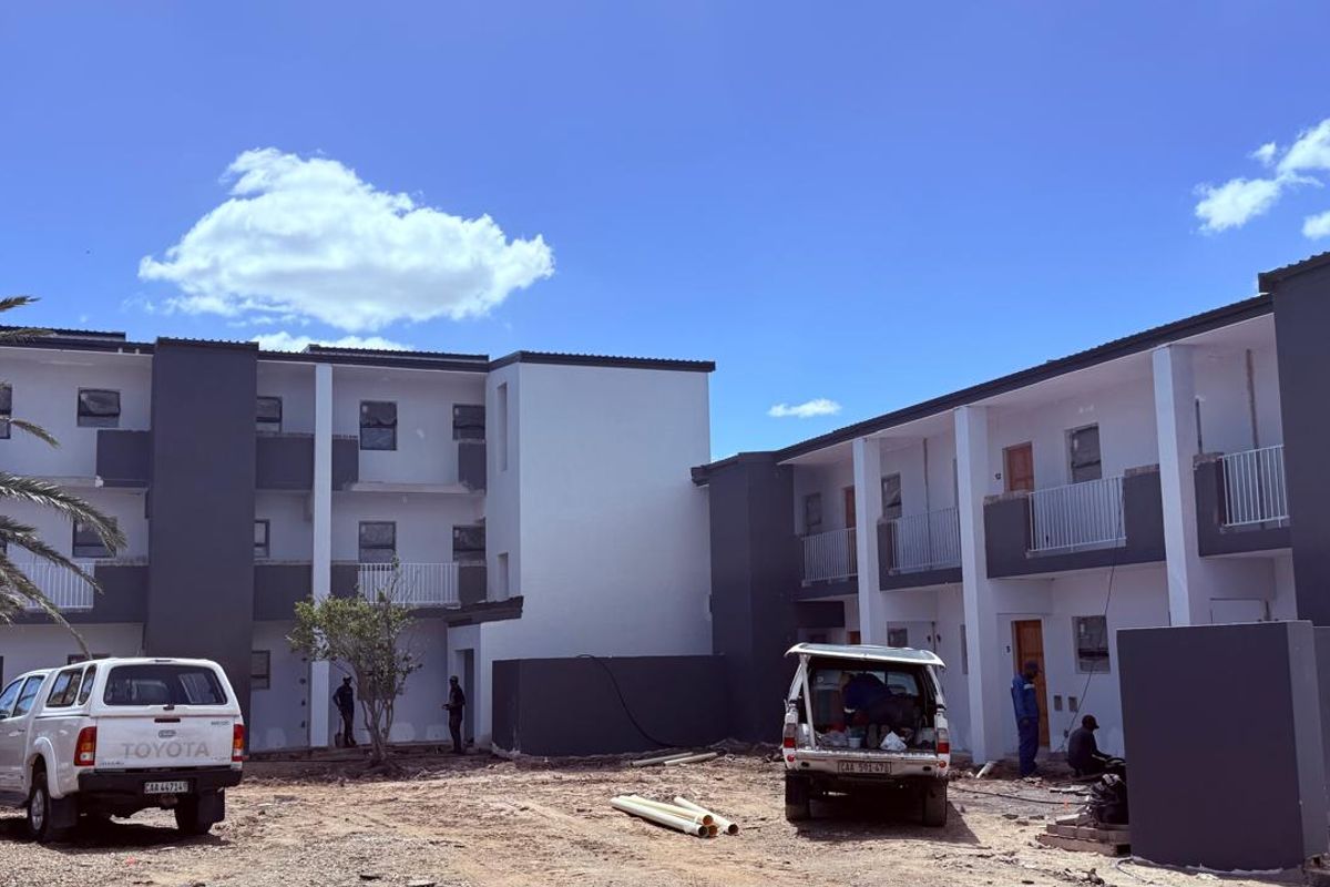 Exterior view of a modern multi-story apartment complex featuring balconies with metal railings, currently undergoing final site landscaping and finishing works.