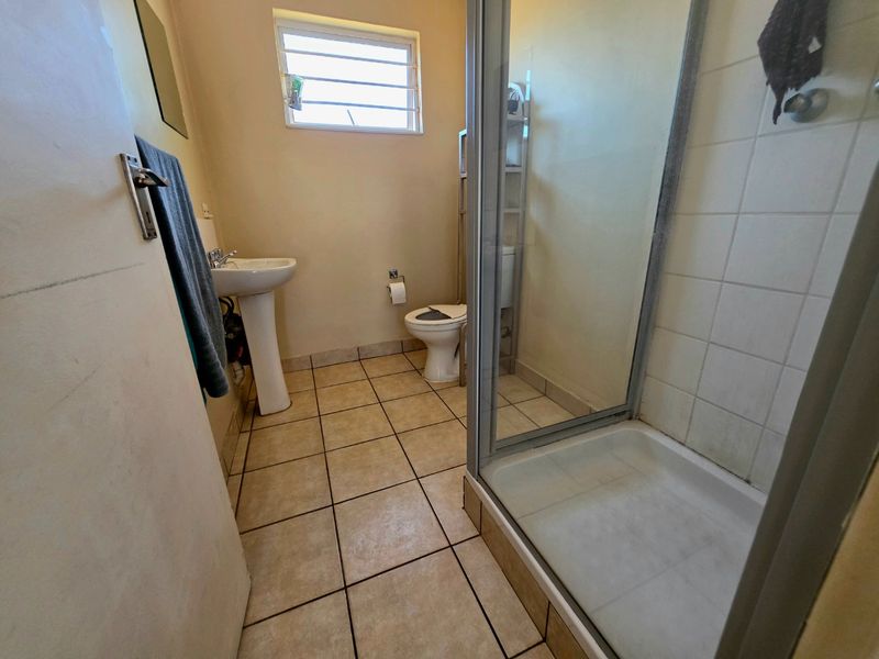 A functional bathroom featuring a pedestal basin, toilet, and a glass-enclosed shower with tiled flooring and a louvered window for ventilation.