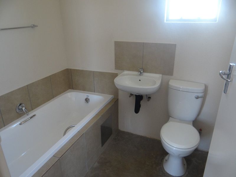A functional bathroom featuring a built-in bathtub, wall-mounted basin, and toilet, with neutral wall tiles and an unfinished concrete floor.
