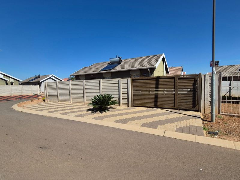 Front exterior view of the property featuring a paved driveway, secure sliding gate, precast boundary wall, and a roof-mounted solar water heating system.