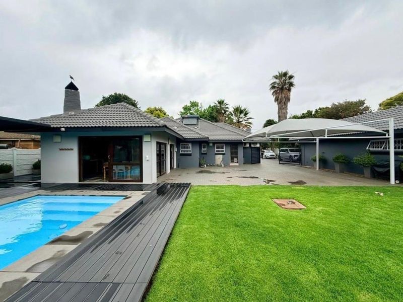 Spacious rear exterior featuring a swimming pool with composite decking, a paved driveway, shade sail, and an outdoor fireplace.