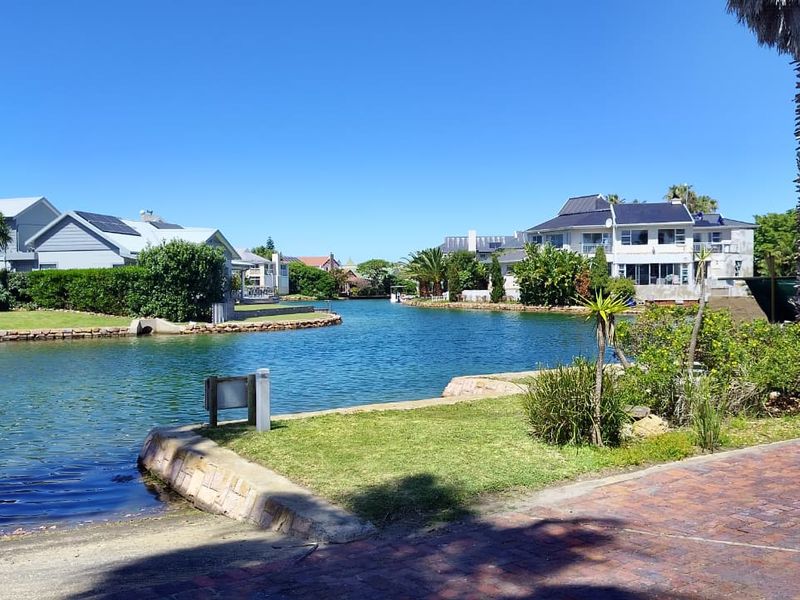 Scenic view of a tranquil canal-front residential area featuring landscaped gardens, stone retaining walls, and paved access.