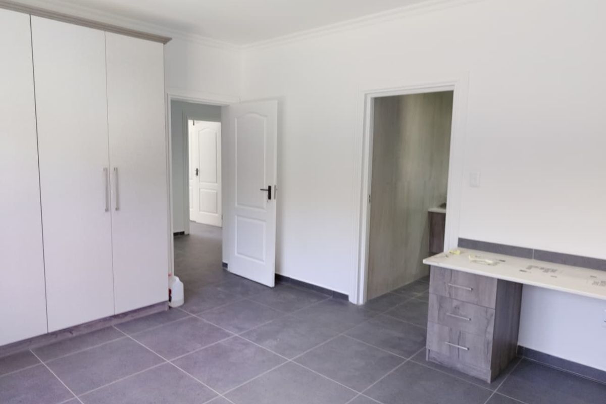 A clean, modern bedroom featuring built-in cupboards, tiled flooring, and a built-in vanity unit, with neutral finishes and direct access to an en-suite bathroom.