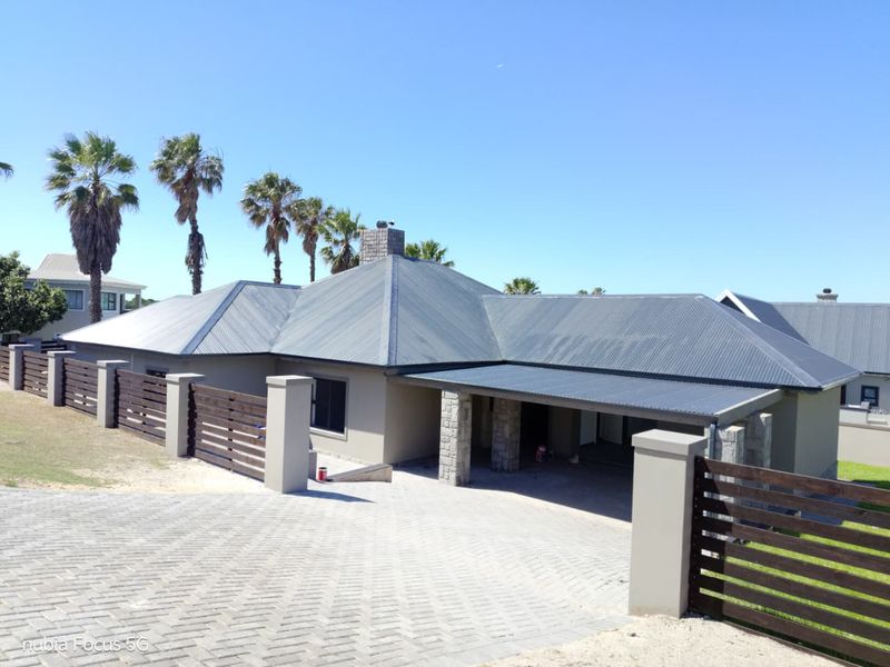 Front exterior view of a modern house featuring a corrugated metal roof, stone-clad pillars, and a paved driveway with timber slat fencing.
