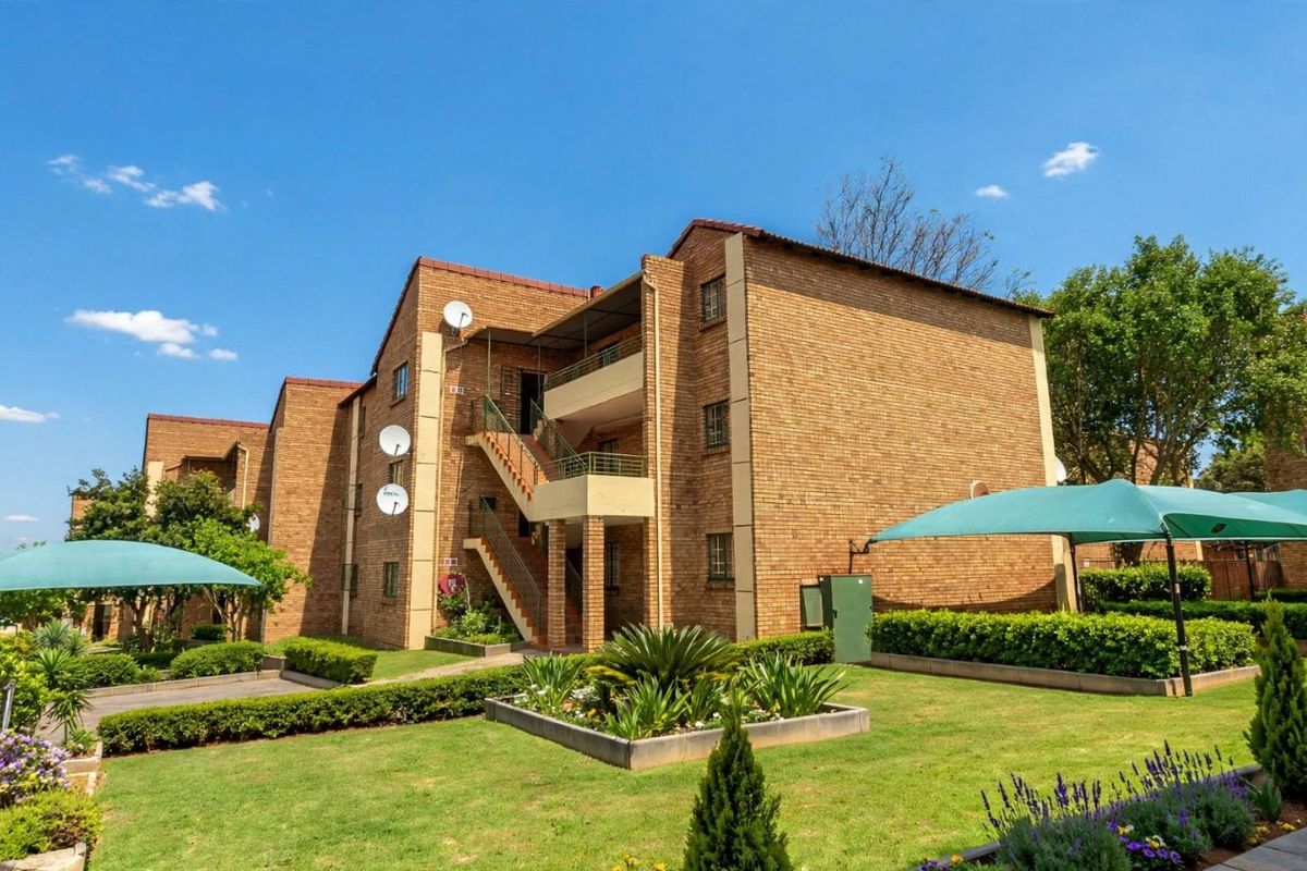 Exterior view of a multi-story apartment complex featuring face-brick architecture, external staircases, balconies, and well-maintained landscaped gardens with shade structures.