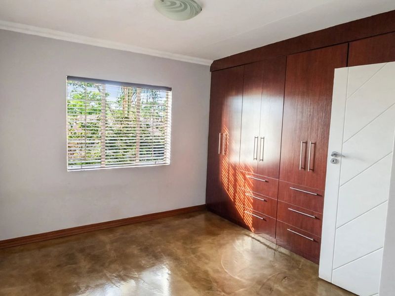 A bright, empty bedroom featuring polished concrete floors, built-in wooden cupboards, and a window with fitted venetian blinds.