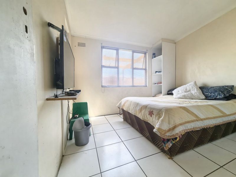 A functional bedroom featuring tiled flooring, built-in shelving, and a window with blinds, offering a basic layout ready for cosmetic updates.