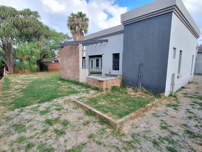 Rear exterior view featuring a paved area, exposed brick feature wall, built-in braai, and timber pergola beams in a garden setting.