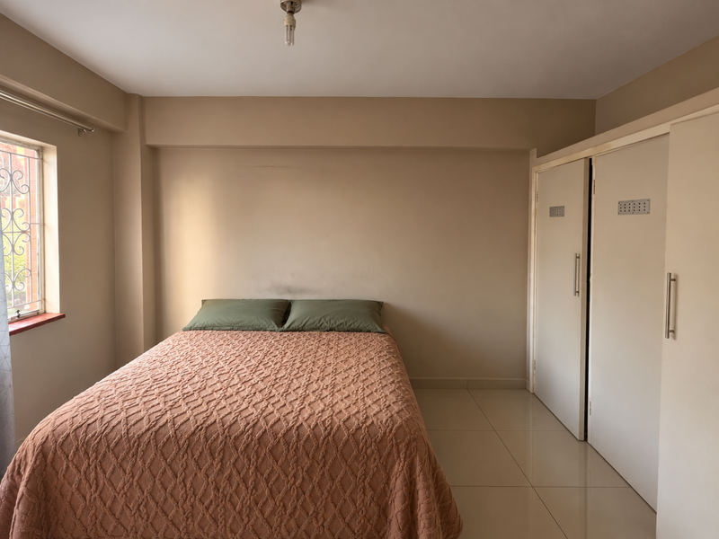 A bedroom featuring neutral wall tones, tiled flooring, and built-in cupboards, offering a functional space with natural light from the side window.
