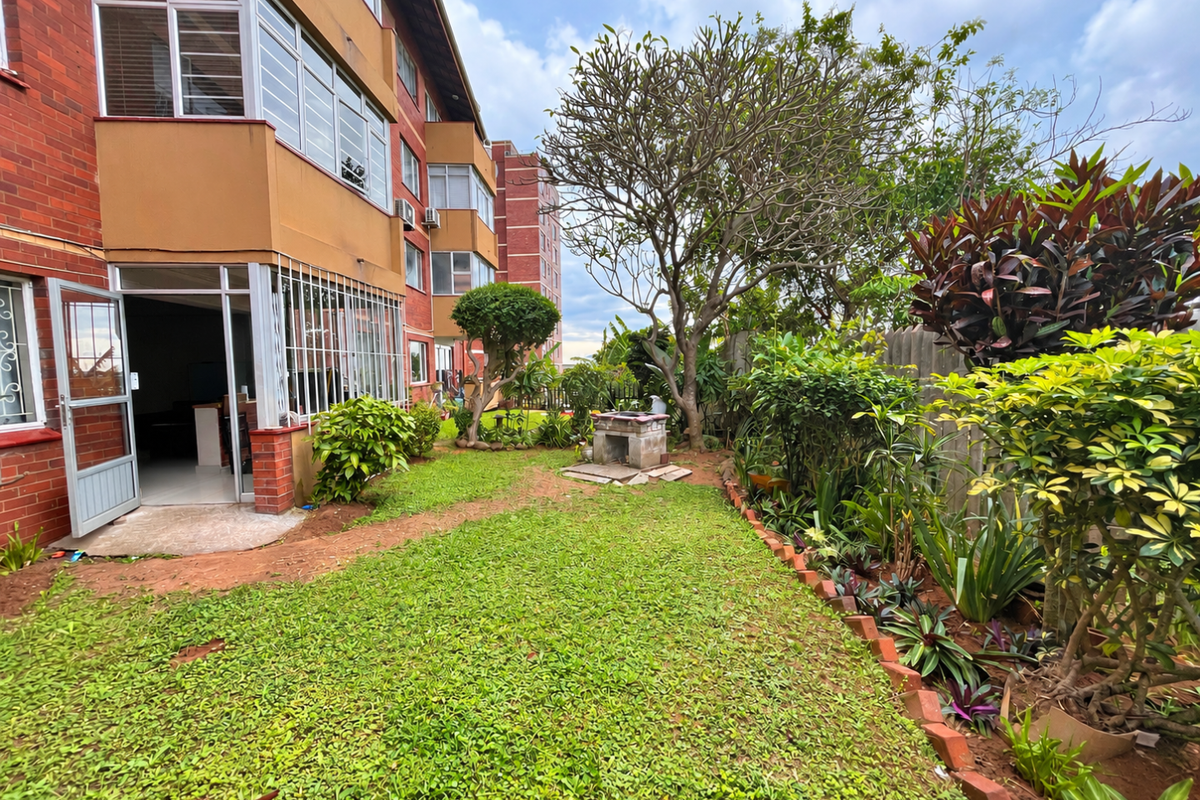 A rear garden view featuring a brick apartment facade, established landscaping, and a built-in brick braai area.