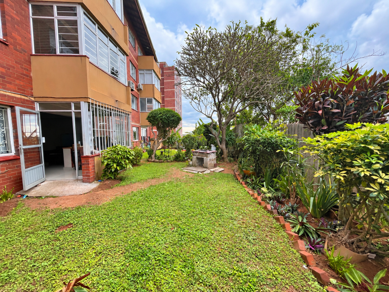 A rear garden view featuring a brick apartment facade, established landscaping, and a built-in brick braai area.