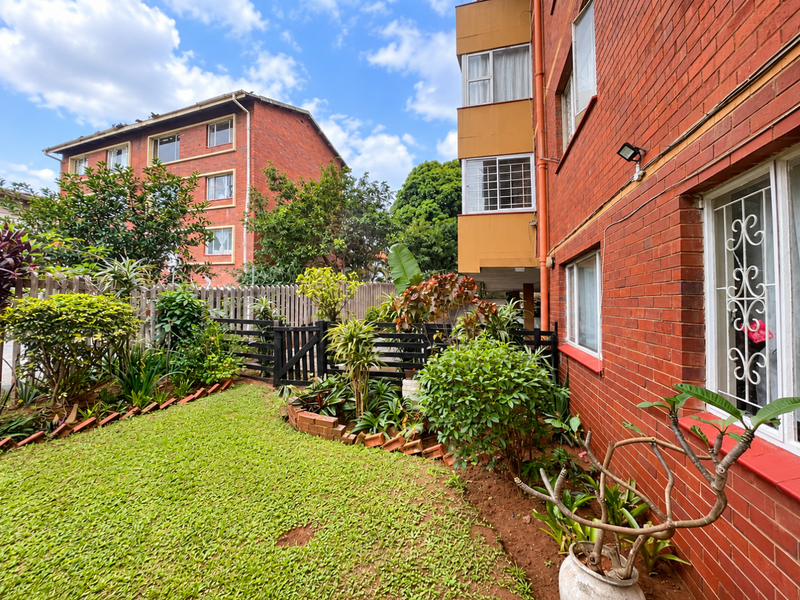 A well-maintained rear garden area featuring a brick apartment facade, security window bars, and established landscaping with a timber boundary fence.