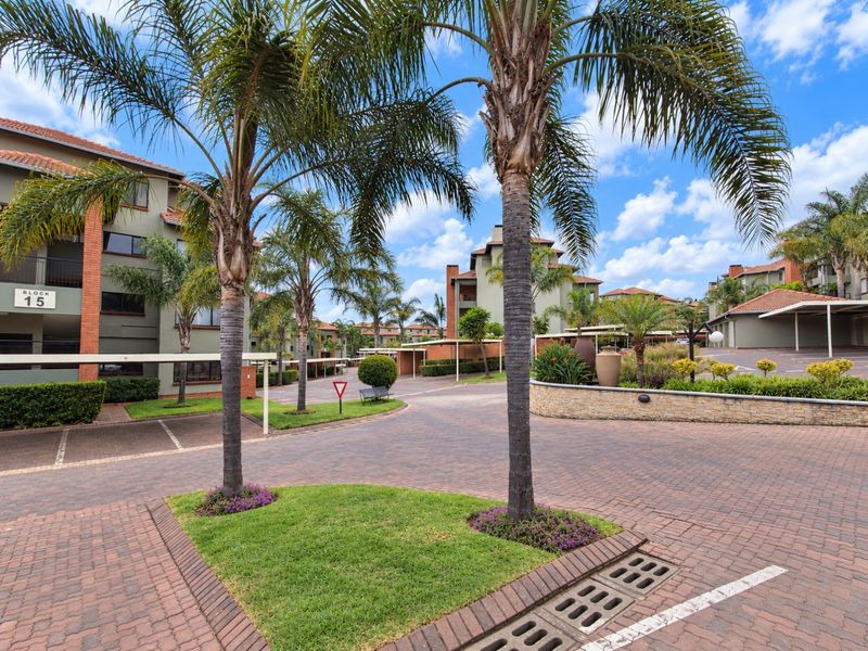 A well-maintained apartment complex exterior featuring paved driveways, landscaped gardens, and covered parking bays within a secure residential estate.