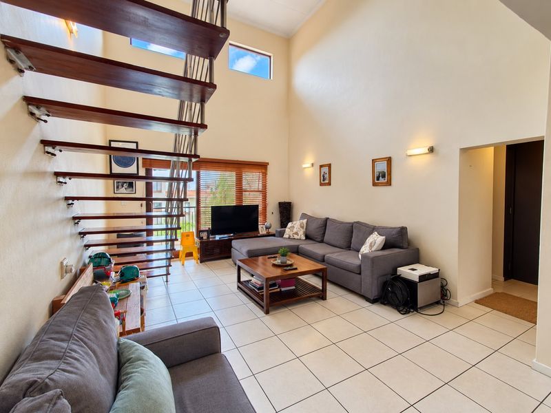 Spacious double-volume living area featuring a modern floating staircase, tiled flooring, and abundant natural light from high-level windows.