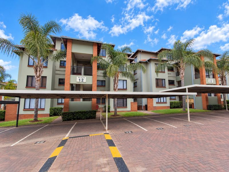 Exterior view of a multi-story apartment complex featuring covered parking bays, paved driveway, and well-maintained landscaped grounds.
