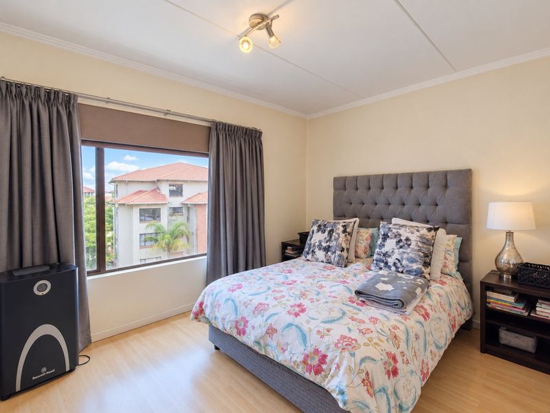 A well-maintained bedroom featuring laminate flooring, neutral wall tones, and a large window providing natural light and views of the surrounding apartment complex.