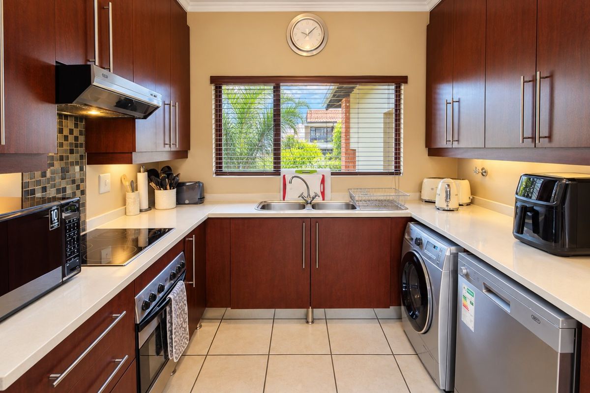 A well-maintained galley kitchen featuring dark wood cabinetry, stone countertops, integrated appliances, and a mosaic tile backsplash with ample under-cabinet lighting.